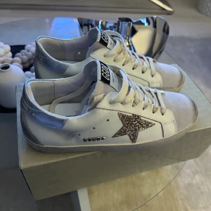 Golden Goose sneakers med glitterstjärna - Hej! säljer ett par snygga vita Golden Goose sneakers med silvriga detaljer. Storlek 39| Lite smuts som går att göra rent enkelt. Kommer med både kvitto, box och ett GGDB sneakers bag till. Säljer billigt- DMa🤩