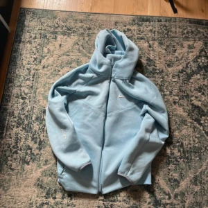 Ljusblå Nike NOCTA hoodiejacka - Säljer en ljusblå hoodiejacka från Nike NOCTA med dragkedja framtill och huva. Jackan har diskret Nike Swoosh på bröstet och NOCTA-logga på ärmen. Tillverkad i mjuk bomullsmix, perfekt för chill dagar. Snygg och clean design med normal passform.