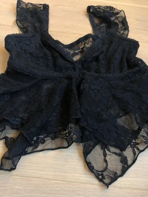 Svart spetsig blus med volang - Säljer en svart blus i transparent spets med blommigt mönster. Toppen har volangdetaljer och är ärmlös med breda axelband.  Jätte fin 