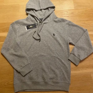 Grå hoodie från Polo Ralph Lauren - Klassisk grå hoodie från Polo Ralph Lauren med svart broderad logga på bröstet. Tröjan har huva med snörning, känguruficka och ribbade muddar. Tillverkad i mjuk bomullsmix som är perfekt för chill dagar.