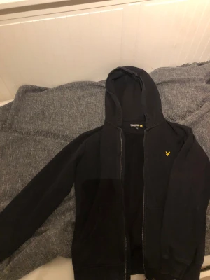 Svart hoodie från Lyle & Scott Junior - Svart hoodie från Lyle & Scott Junior med gul broderad logga på bröstet. Tröjan har huva, dragkedja framtill och två fickor. Materialet är mjuk bomull och modellen är normal passform med långa ärmar. Perfekt för chill eller häng med kompisar.