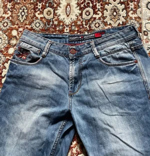 Blå jeans från Tommy Hilfiger - Snygga blå jeans från Tommy Hilfiger med klassisk femficksdesign och coola röda detaljer på knapp och insida. Jeansen har en rak passform och subtila slitningar för en avslappnad vibe. Perfekt för dig som gillar stilrena och tidlösa jeans. Innerbenet: 80cm ytterben: 105cm skrev: 25 midja: 42