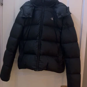 Svart pufferjacka från Calvin Klein Jeans - Svart pufferjacka från Calvin Klein Jeans med huva och dragkedja framtill. Jackan är vadderad med dun, har diskret CK-logo på bröstet och är tillverkad av återvunnen nylon och polyester. Perfekt för kalla dagar och har en clean, modern look.