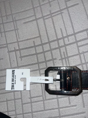 Svart skärp True Religion skinn M/L - Säljer ett svart bälte från True Religion i skinn med coolt silverfärgat spänne där loggan är ingraverad. Bältet har röda kontrastsömmar som ger en snygg detalj och en modern vibe. Perfekt för att lyfta din outfit med en exklusiv touch.