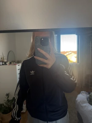 Marinblå Adidas bomberjacka - Säljer en marinblå Adidas bomberjacka med klassiska vita ränder längs ärmarna och Adidas-logga på bröstet. Jackan har dragkedja framtill, ribbade muddar och är långärmad. Perfekt för en sportig och chill stil.