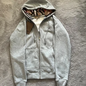 Grå Burberry hoodie med rutig huva - Snygg grå hoodie från Burberry med klassiskt rutigt mönster i huvan och insidan. Tröjan har dragkedja, snörning vid huvan och broderad Burberry-logga på bröstet. Mjukt material och två fickor framtill. Perfekt för dig som gillar stilrena detaljer.