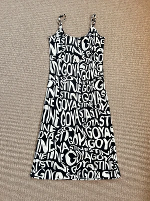 Svartvit långklänning Stine Goya - Cool långklänning från Stine Goya med svart botten och vit grafisk text över hela klänningen. Klänningen har smala axelband och en rak, något utställd siluett. Materialet är mjukt och följsamt, perfekt för dig som gillar statement-plagg med unik design.