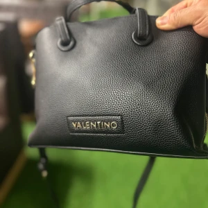 Svart handväska från Valentino - Snygg svart handväska från Valentino med guldiga detaljer och präglad yta. Väskan har både handtag och axelrem, samt dragkedja med Valentino-logga. Insidan är fodrad och har en innerficka med dragkedja. Perfekt storlek för det viktigaste.