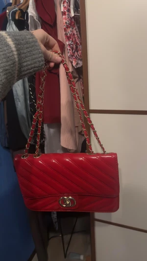 Röd quiltad axelväska med kedja - Snygg röd axelväska med quiltad design och guldfärgad kedja som axelrem. Väskan har ett vridlås i guld och är tillverkad i glansigt skinnliknande material. Perfekt för att lyfta din outfit med en pop av färg och coola detaljer.