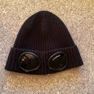 Svart ribbad mössa C.P. Company goggles - Svart ribbstickad mössa från C.P. Company med ikoniska goggles framtill på uppviket. Mössan är tillverkad i mjuk ull och har en klassisk form med bred uppvikt kant. Perfekt accessoar för att sticka ut i vinterkylan.