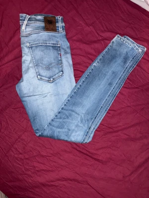 Replay Anbass ljusblå jeans, strl 30 - Säljer ett par Replay Anbass jeans i ljusblå tvätt med snygga slitningar och klassisk femficksdesign. Jeansen har smal passform, normal midja och är tillverkade i mjukt jeansmaterial med stretch. Perfekt för dig som gillar en modern och avslappnad look.