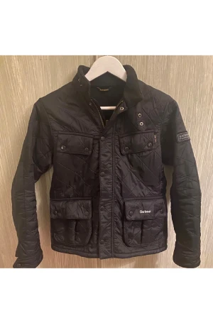 Svart quiltad jacka från Barbour - Quiltad svart jacka från Barbour med ståkrage, flera fickor med lock och tryckknappar, samt Barbour International patch på ärmen. Jackan har dragkedja och knappar framtill och är tillverkad i syntetmaterial. Perfekt för dig som gillar klassisk stil med edge.