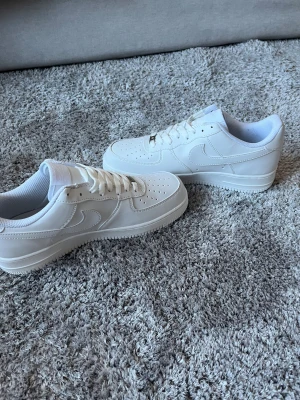 Nike Air Force 1 helvita sneakers - Nike Air Force 1 sneakers i helvit färg med klassisk låg siluett. Skorna har snörning, perforerad tå och rundad sula. Tillverkade i skinn med vit mesh på insidan för extra komfort. Snyggt broderad Nike Air-logga på hälen och metallplatta på snörningen.