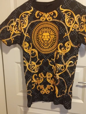 Svart t-shirt med lejonmotiv från Icono - Svart t-shirt från Icono med slim fit-passning och korta ärmar. T-shirten har ett stort lejonmotiv i guld och detaljerade barockinspirerade mönster i gult och guld över hela plagget. Materialet är mjuk bomull och trycket är riktigt iögonfallande.