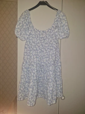 Blommig ljusblå klänning Gina Tricot XS - Supersöt ljusblå kortklänning från Gina Tricot i storlek XS. Klänningen har puffiga kortärmar, v-ringning med knyt framtill och ett vitt blommigt mönster. Tillverkad i mjuk bomull, perfekt för sommarens alla tillfällen. Volangdetaljer ger en extra feminin touch.