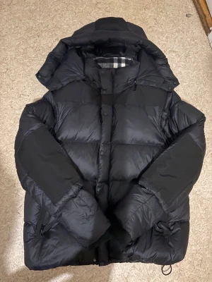 Svart pufferjacka Burberry - Svart pufferjacka från Burberry med klassiskt rutigt foder i kragen. Jackan har en stor huva, dragkedja och knappar framtill samt praktiska fickor med dragkedja. Materialet är polyester och dun, perfekt för kalla dagar. Snygg och modern look med loose passform.