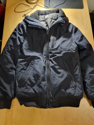 Svart teddyfodrad pufferjacka FSBN XS - Svart pufferjacka från FSBN i storlek XS med teddyfoder i hela insidan och huvan. Jackan har dragkedja framtill, flera fickor med dragkedja och ribbade muddar vid ärmslut och nederkant. Perfekt för kalla dagar med sin tjocka och mysiga känsla.