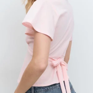 Ljusrosa omlottopp från Gina Tricot - Supersöt ljusrosa omlottopp från Gina Tricot med v-ringning och korta, lite fladdriga ärmar. Toppen har knytband i midjan som ger en snygg siluett. Perfekt att styla med jeans eller kjol för en fräsch look.