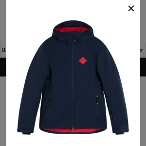 Mörkblå skidjacka från J.Lindeberg   - Snygg mörkblå skidjacka med röd insida och röd logga på bröstet. Jackan har huva, hel dragkedja framtill och sidoficka med dragkedja. Perfekt för blåsiga dagar och skidåkning, enkel att matcha med det mesta. Modellen starling. Nypris ca 4000kr 