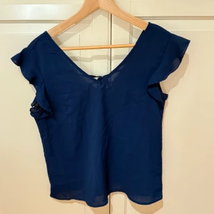 Marinblå blus med spets och volang - Snygg marinblå blus med v-ringning och spetsdetaljer vid halsen. Blusen har korta volangärmar och ett knyt i ryggen som ger en extra touch. 