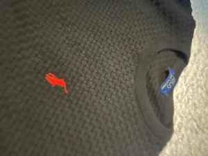 Svart stickad tröja från Polo Ralph Lauren - Svart stickad tröja från Polo Ralph Lauren med klassisk rund hals och ribbade muddar. Tröjan har ett snyggt vävt mönster och den ikoniska röda Polo-loggan broderad på bröstet. Perfekt för dig som gillar stilrena och tidlösa plagg.
