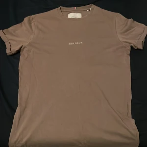 Beige t-shirt från Les Deux XS - Snygg beige t-shirt från Les Deux i storlek XS. T-shirten har en klassisk rund hals och korta ärmar. På bröstet finns en stilren vit Les Deux-logga. Materialet är mjuk bomull som känns skönt mot huden. Perfekt för dig som gillar minimalistisk och clean stil.
