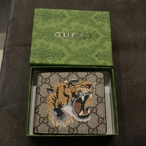 Gucci plånbok med tigerprint - Snygg plånbok från Gucci med klassiskt GG-mönster och en fet tiger på framsidan. Insidan har flera kortfack och fönster för ID, med röd Gucci-logga. Materialet är skinn och färgerna går i beige, brun och svart med detaljer i orange och vit.