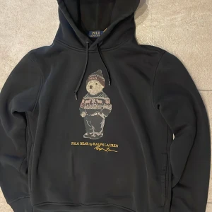 Svart hoodie Polo Bear Ralph Lauren - Svart hoodie från Polo Ralph Lauren med ikoniska Polo Bear-trycket på bröstet. Tröjan har snörning i huvan och är tillverkad i mjuk bomull och polyester. Perfekt för dig som gillar streetwear och vill sticka ut med en cool print.