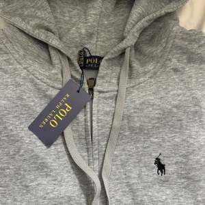 Grå hoodie från Polo Ralph Lauren - Snygg grå hoodie från Polo Ralph Lauren med klassisk broderad logga på bröstet. Tröjan har huva med snörning och dragkedja framtill. Mjuk bomullskvalitet och stilren look som funkar till jeans eller joggers. Perfekt för chill eller när du vill vara casual.