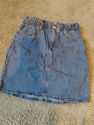 Blå jeanskjol från Lindex - Säljer en klassisk blå jeanskjol från Lindex med resår i midjan. Kjolen har fickor både fram och bak samt hällor för bälte. Perfekt för dig som gillar en enkel och tidlös denimstil.