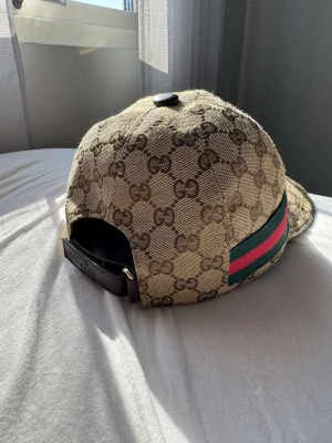 Gucci keps med GG-mönster - Snygg beige keps från Gucci med klassiskt GG-monogram över hela kepsen. Har en grön och röd rand på sidan och justerbar rem med läderdetalj baktill. Perfekt för dig som vill ha en lyxig och trendig accessoar.