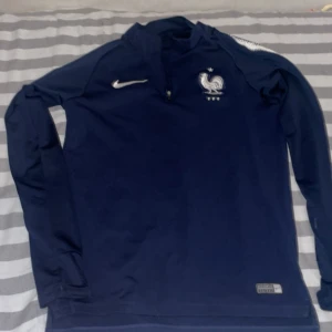 Blå långärmad Nike Frankrike-tröja - Säljer en mörkblå långärmad fotbollströja från Nike med Frankrikes landslagsemblem och FFF-logga på bröstet. Tröjan har halv dragkedja framtill och är tillverkad i ett sportigt, lätt material som andas. Perfekt för träning eller match!