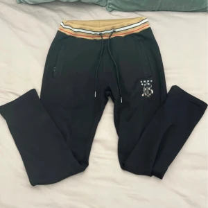 Svarta Burberry mjukisbyxor strl xs-S - Svarta mjukisbyxor från Burberry med klassisk TB-logga i rutigt mönster på benet. Byxorna har beige, vit och röd randig resår i midjan samt snörning. Mjukt material och fickor med dragkedja framtill. Perfekta för en chill och stilren look.