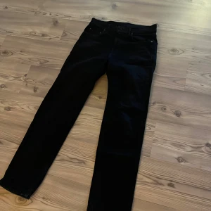 Svarta jeans från Dressman  - Svarta jeans från Dressman med raka ben. Jeansen har bälteshällor och en snygg läderpatch med logga bak i midjan. Materialet är slitstarkt jeans och passformen är normal, perfekt för dig som gillar stilrena svarta jeans.