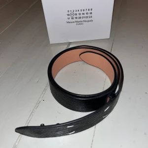 Maison margiela bälte - Maison margiela bälte i genuine leather. Svart med four stitch loggan där framme.  Bältet var lite för kort för mig o därför säljer jag. Knopparna är lite slitna där de sitter fast men det är inget man märker när bältet är på eftersom att de pekar inåt. Köpt i London i butik. Har påsen men hittade inte själva kvittot när jag packade upp min väska, finns dock en lapp och ett kort i boxen. Kommer med originalförpackning och dustbag.              Pris kan diskuteras och skickas snabbt.