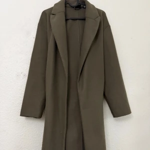 Olivgrön kappa från Vero Moda - Snygg olivgrön kappa från Vero Moda med klassisk krage och knytbälte i midjan. Jackan har lång ärm och rak passform, perfekt för dig som gillar stilrena och enkla plagg. Materialet är mjukt och känns skönt mot huden.