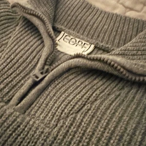Grå stickad half zip-tröja COPE - Snygg grå stickad tröja från COPE med ribbad struktur och half zip-dragkedja vid kragen. Tröjan har långärmad passform och en klassisk, lite loose siluett som är perfekt för kyliga dagar. Enkel att matcha med jeans eller cargo för en chill look.