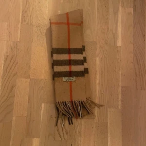 Beige rutig halsduk från Burberry - Klassisk beige halsduk från Burberry i 100% kashmir med rutigt mönster i brunt, svart och rött. Tidlös design med fransar i ändarna och den ikoniska Burberry-taggen. Perfekt accessoar för att lyfta din outfit.