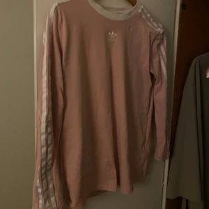 Rosa långärmad Adidas tröja - Säljer en snygg rosa långärmad tröja från Adidas med klassiska vita ränder längs ärmarna och broderad logga på bröstet. Tröjan har ribbad vit krage och är gjord i mjuk bomull.