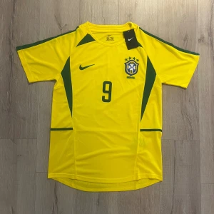 Brasilien Ronaldo #9 fotbollströja Nike - Gul Brasilien fotbollströja från Nike med gröna detaljer och nummer 9 på bröstet och ryggen. RONALDO står tryckt på ryggen. Tröjan har korta ärmar, rund hals och är tillverkad i lätt polyester. CBF-emblem broderat på framsidan och Nike-logga. Perfekt för fans av brasiliansk fotboll.