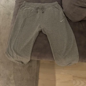 Grå mjukisbyxor från Nike, storlek L - Grå mjukisbyxor från Nike i storlek L med klassisk logga på vänster ben. Byxorna har snörning i midjan, ribbade muddar vid benslut och är tillverkade i mjukt bomullsmaterial. Perfekta för chill eller träning, med två sidofickor och relaxed passform.