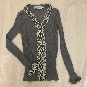 Grå kofta med volang från Anne Fontaine - Supercool unik grå ribbad kofta med vita volangdetaljer framtill. Långärmad och figurnära modell som ger en chic och trendig look. Ge gärna prisförslag