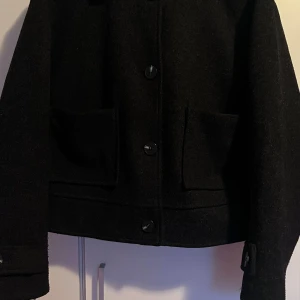Svart ulljacka från 157, storlek S - Svart jacka från 157 i ullmaterial med klassisk rak passform. Jackan har stora fickor framtill, knäppning med tre knappar och detaljer vid ärmslut. Perfekt för dig som gillar stilrena och enkla plagg med lite edge.