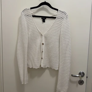 Vit stickad kofta från Lindex - Säljer en vit stickad kofta från Lindex i storlek M. Koftan har ett luftigt hålmönster, v-ringad hals och snygga knappar framtill. Långärmad och med ribbade muddar vid ärmslut och nederkant. Perfekt för lager-på-lager under vår och sommar.