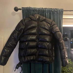 Svart pufferjacka från Colmar - Svart pufferjacka från Colmar med glansig finish, huva och två fickor med dragkedja. Jackan har ett snyggt Colmar-märke på ärmen och är perfekt för kalla dagar. Modellen är quiltad och har en normal passform med lång ärm. Jackan är i storlek 152 och funkar som xxs-xs. Köpt på kidsbrandstore för 4000