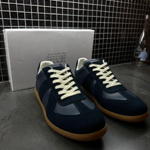 Maison Margiela sneakers marinblå mocka - Maison Margiela sneakers i marinblå mocka och skinn med vita snören och klassisk gummisula. Snygg retrostil med rund tå och kontrasterande detaljer. Perfekta för dig som gillar stilrena och exklusiva sneakers med streetkänsla.