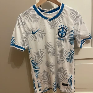 Vit Brasil Nike fotbollströja med blå detaljer - Vit Brasilien fotbollströja från Nike med blå logga och emblem. Tröjan har grå palmbladsmönster över hela och blå detaljer vid krage och ärmslut. Broderat CBF-märke på bröstet och klassisk Nike swoosh. Perfekt för dig som älskar unik design.