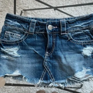 Blå jeanskjolmed slitningar, snygg bakficka - !!Priset kan diskuteras!! Använd 2 gånger                                 Snygg blå jeanskjol med slitna detaljer och fransig kant. Klassisk femficksmodell med vita kontrastsömmar och bälteshällor. Perfekta för sommaren och ger en cool, avslappnad vibe.