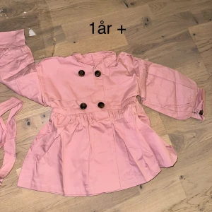 Rosa trenchcoat med knappar, barn. Kappa  - Söt rosa trenchcoat för barn med dubbelknäppning och volangdetaljer vid midjan. Jackan har långa ärmar, klassisk krage och medföljande bälte i samma färg. Perfekt för dig som gillar gulliga och stilrena plagg till de små.Söt rosa kappa med knappar och skärp.  🎁 