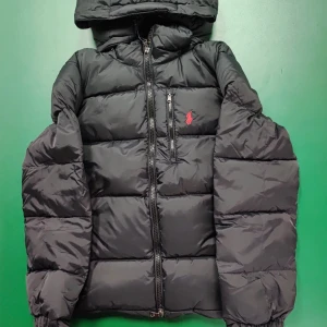 Svart pufferjacka från Polo Ralph Lauren - Svart pufferjacka från Polo Ralph Lauren med huva och röd logga på bröstet. Jackan har en quiltad design, dragkedja framtill och en bröstficka med dragkedja. Perfekt för kalla dagar och har en riktigt snygg streetstil. Kom med prisförslag